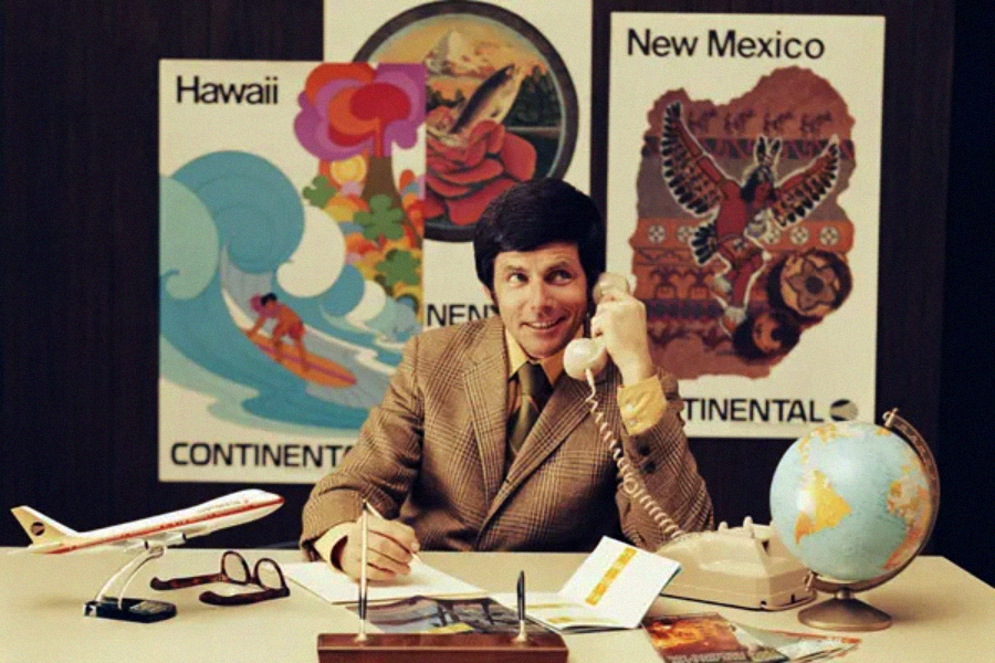 A man in a brown plaid suit sits at a desk with a telephone, globe, and travel brochures. Behind him are colorful vintage travel posters for Hawaii and New Mexico. A model airplane and glasses are also on the desk.