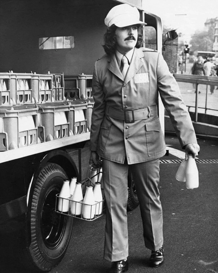 A person in a uniform and cap carries a metal crate filled with glass milk bottles, walking beside a delivery truck loaded with more bottles. The street scene appears to be from a past era, likely the 1970s.