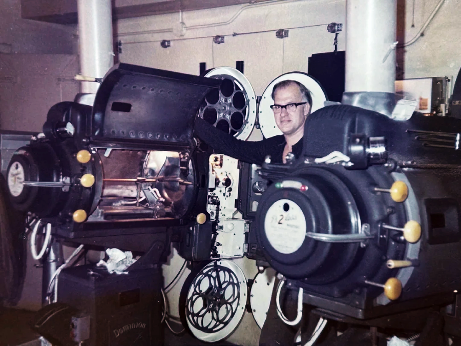A man stands between two large vintage film projectors in a projection room, with various film reels and equipment visible around him. One projector door is open, showing the film mechanism inside.