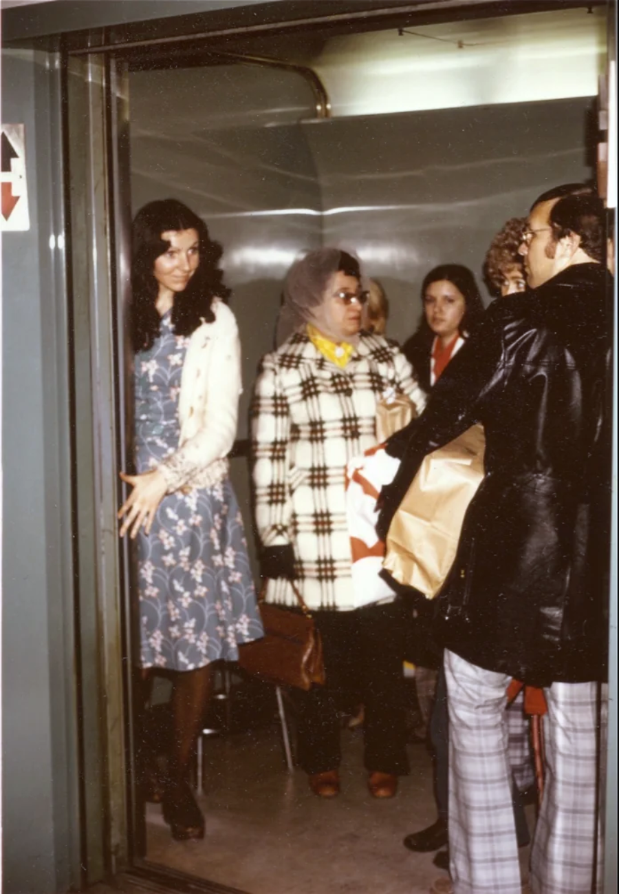 A group of people, including women in patterned dresses and coats, stand inside an open elevator. A woman in a floral dress stands at the door, while others wait outside, one holding a large wrapped package.