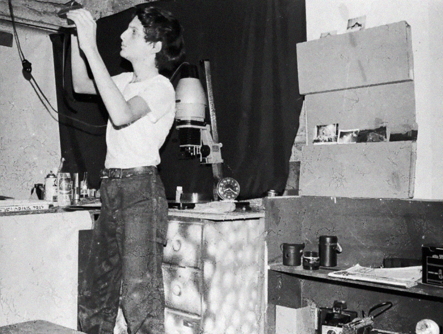 A person in a white shirt and jeans works in a home photo darkroom, handling photographic film near an enlarger. Shelves hold mugs, papers, and photography equipment. A dark cloth hangs behind the workspace.