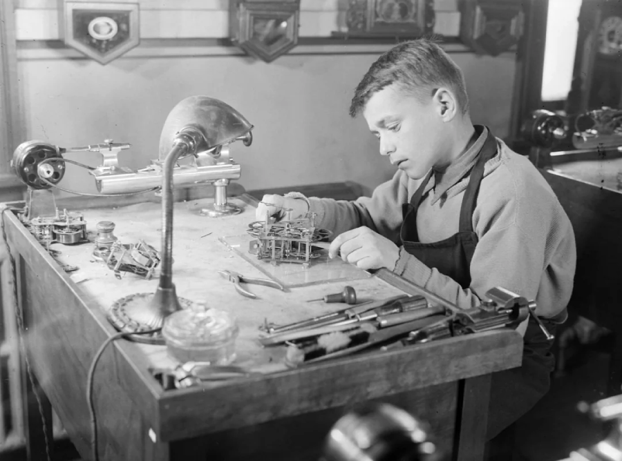 A boy sits at a workbench, focused on assembling clock parts. Various tools and clock components are spread out on the table, and a desk lamp illuminates his workspace.
