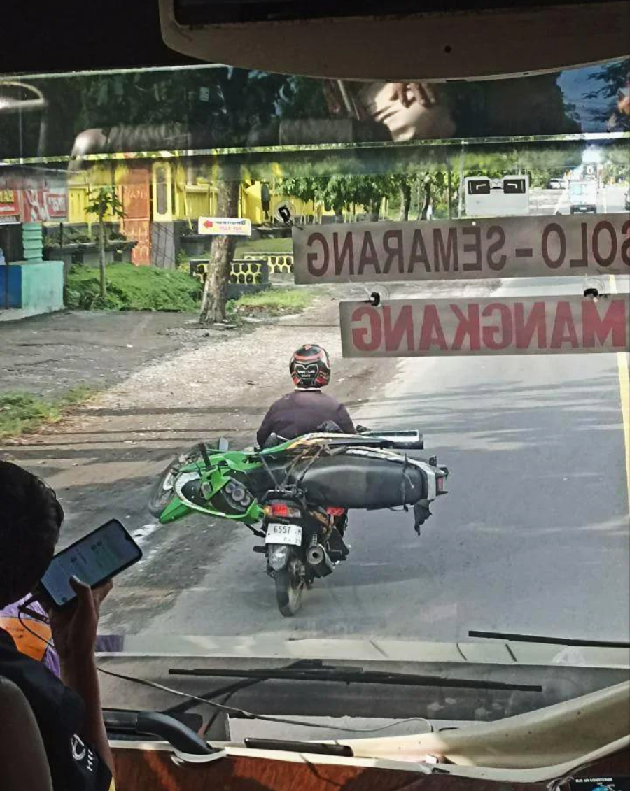 A person rides a motorcycle carrying a green bicycle sideways on the back, seen from inside a bus. A passenger in the foreground looks at their phone. The road is lined with trees and buildings.