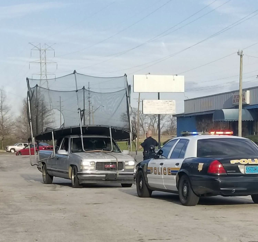 A silver car is stopped by a police car, with a large trampoline, including its net, awkwardly strapped on top and hanging over the sides. Two people stand nearby on a paved lot.