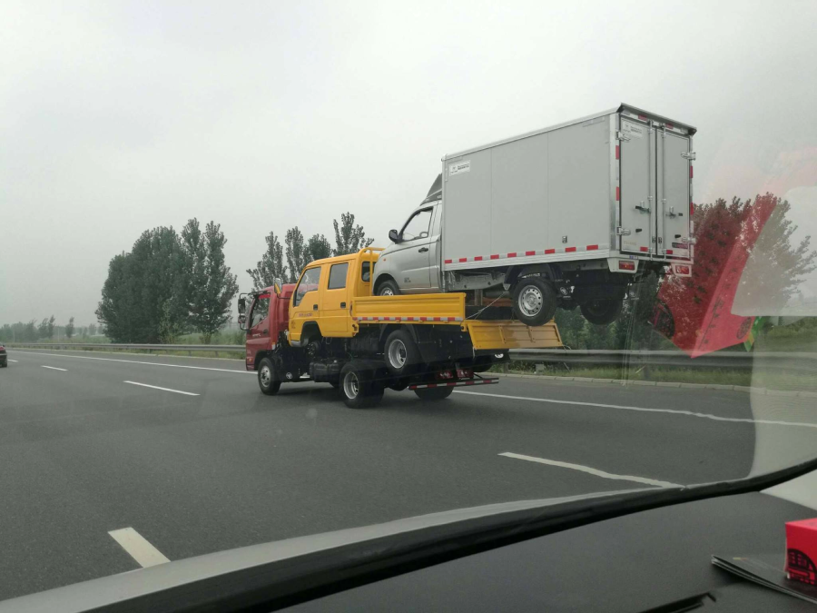 A red tow truck carries a yellow flatbed truck, which is also carrying a large silver delivery van, all stacked on top of each other while driving on a multi-lane highway. Trees line the road under a cloudy sky.