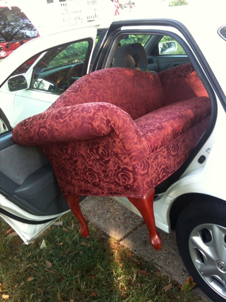 A large, red-patterned couch is awkwardly wedged into the backseat of a white car, with the door open and the legs sticking out, showing an attempt to transport furniture in a small vehicle.