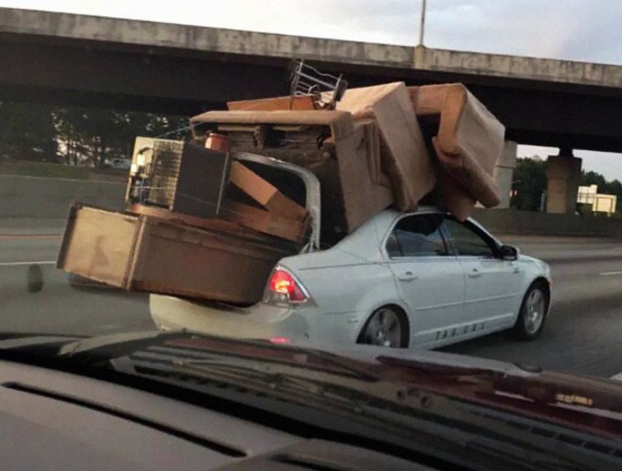 A white sedan driving on a highway, overloaded with large furniture items like a sofa, dresser, and chairs stacked on and protruding from the trunk and roof, with its trunk unable to close.