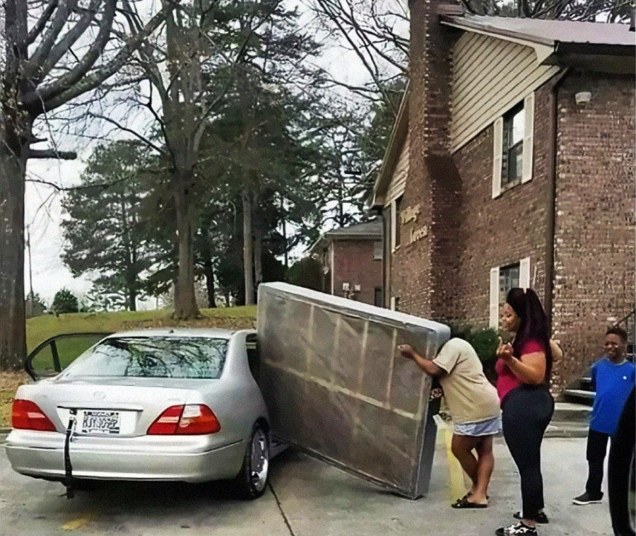 Three people struggle to fit a large mattress into the trunk of a silver car outside an apartment building, while a woman and a child watch and laugh nearby.