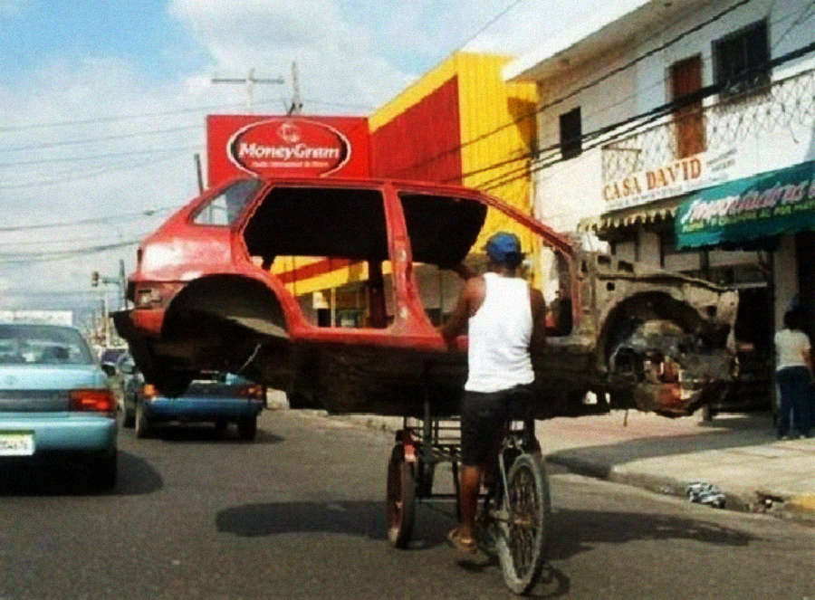 A man pedals a tricycle cart carrying the stripped shell of a red car down a busy street, passing shops and parked vehicles.