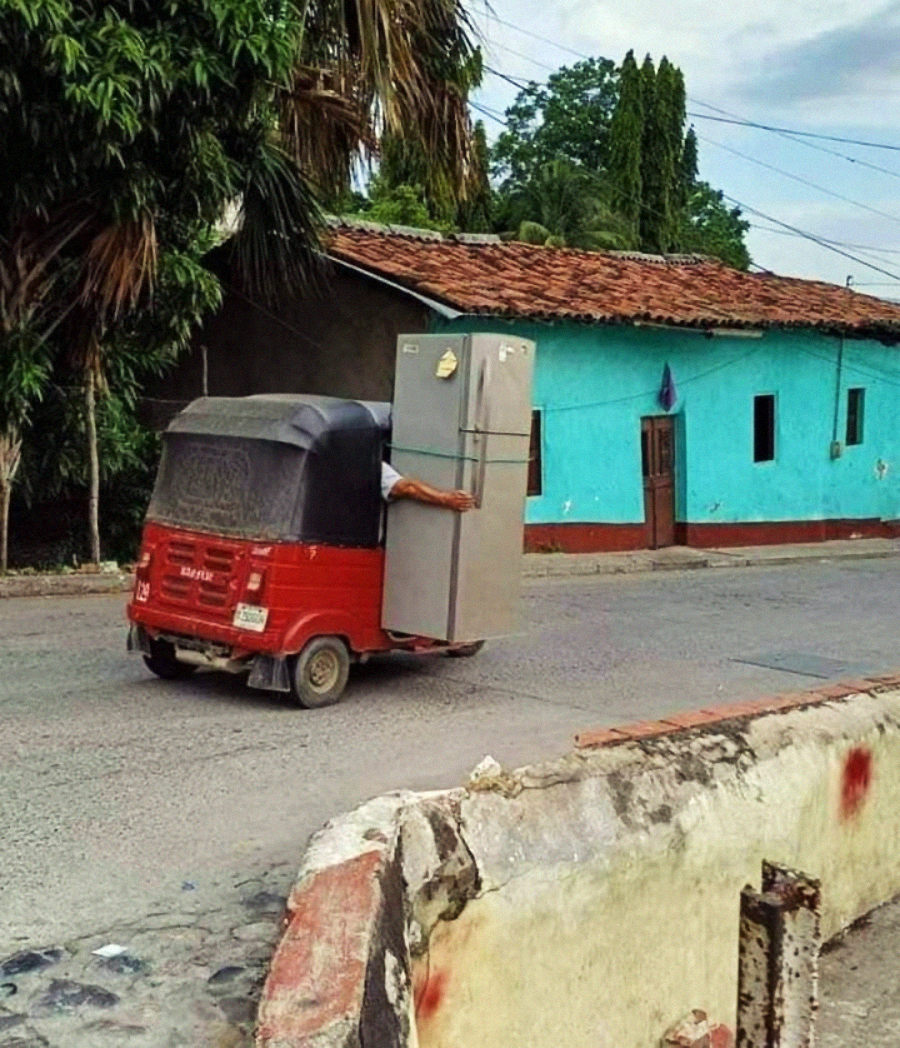 A red auto rickshaw drives down a street, transporting a large refrigerator with someone’s arm visible reaching out to hold it upright, passing by a turquoise house with a tiled roof.