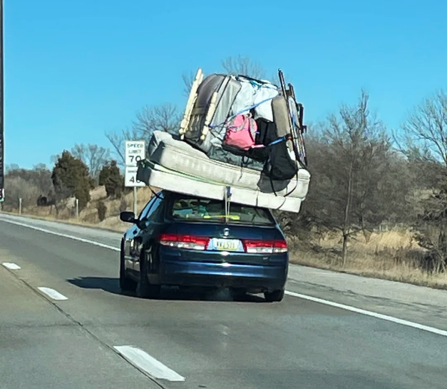 A car drives down the highway with mattresses, furniture, and various household items precariously stacked and tied to its roof. The sky is clear and the surrounding area is rural with dry grass and some trees.