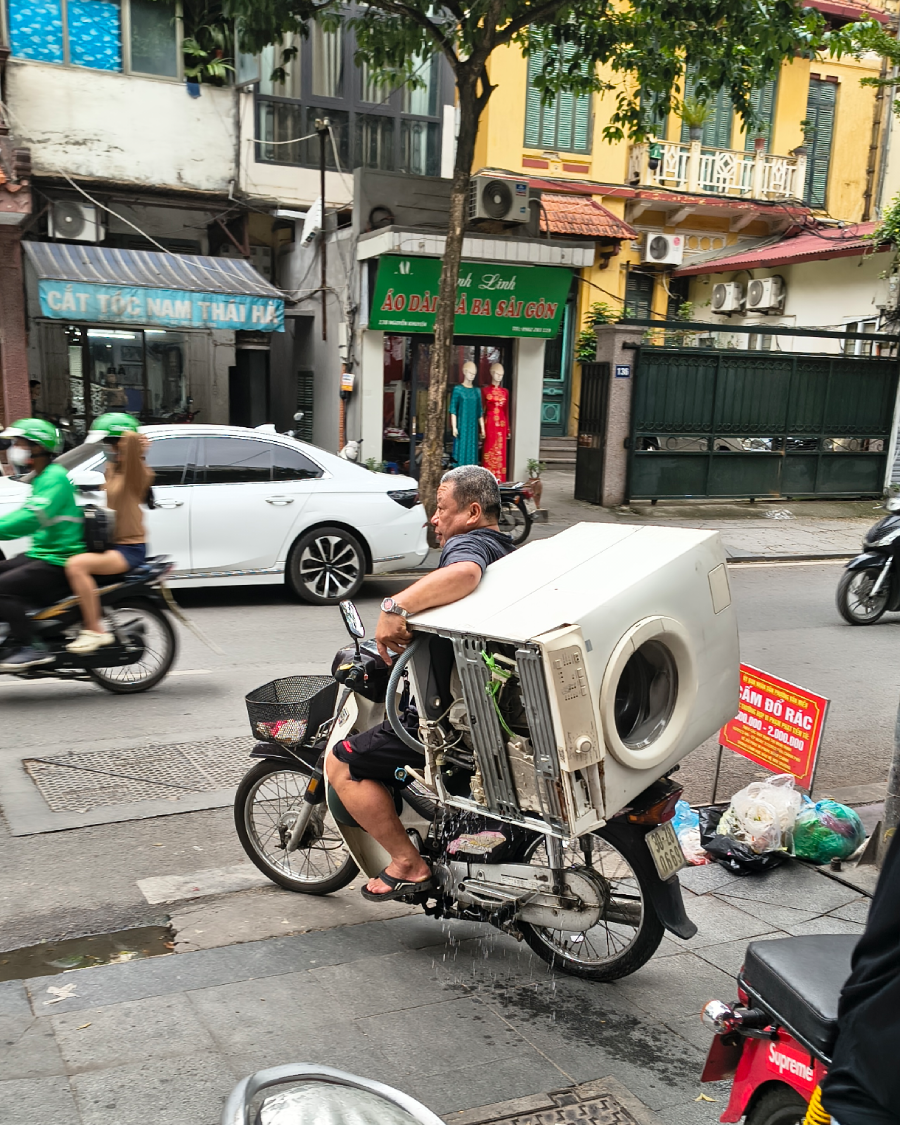 A man rides a motorbike through a busy city street carrying a large washing machine strapped to his back, with cars and motorbikes passing by in the background.