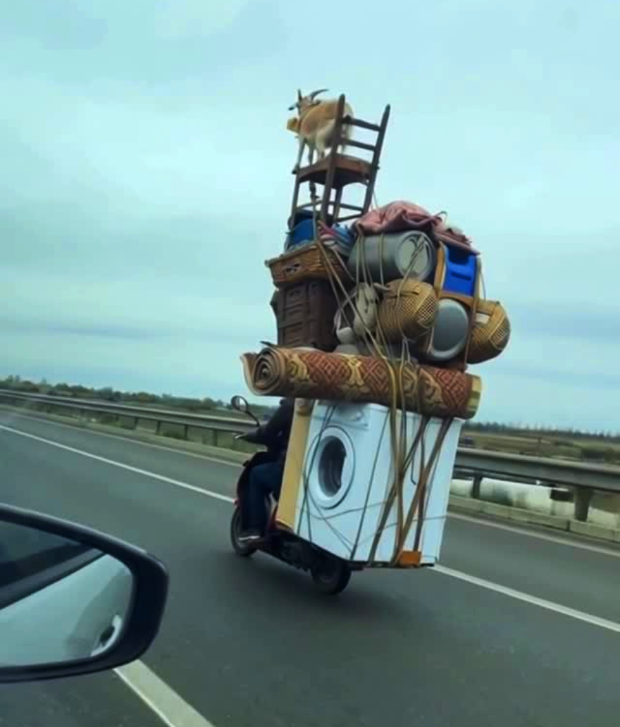 A person rides a motorcycle on a highway, carrying an enormous, precarious stack of household items including a washing machine, chair, baskets, rugs, and other belongings tied together.