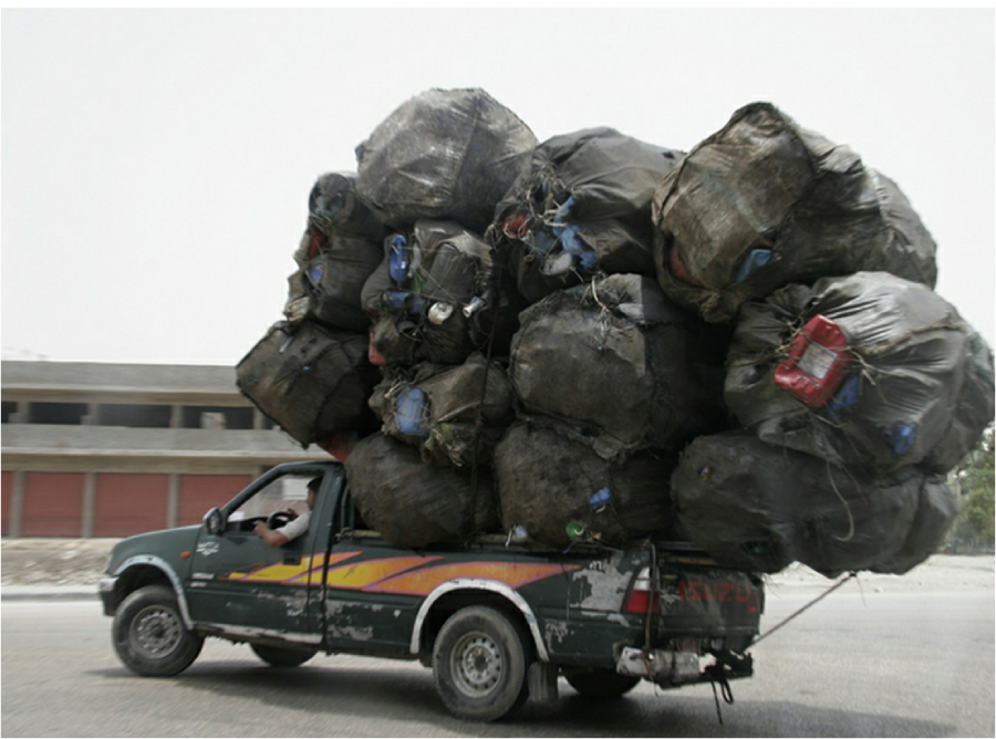 A small green pickup truck drives down a road overloaded with large black bags and bundles stacked high and tied together, causing the vehicle to sag under the weight.