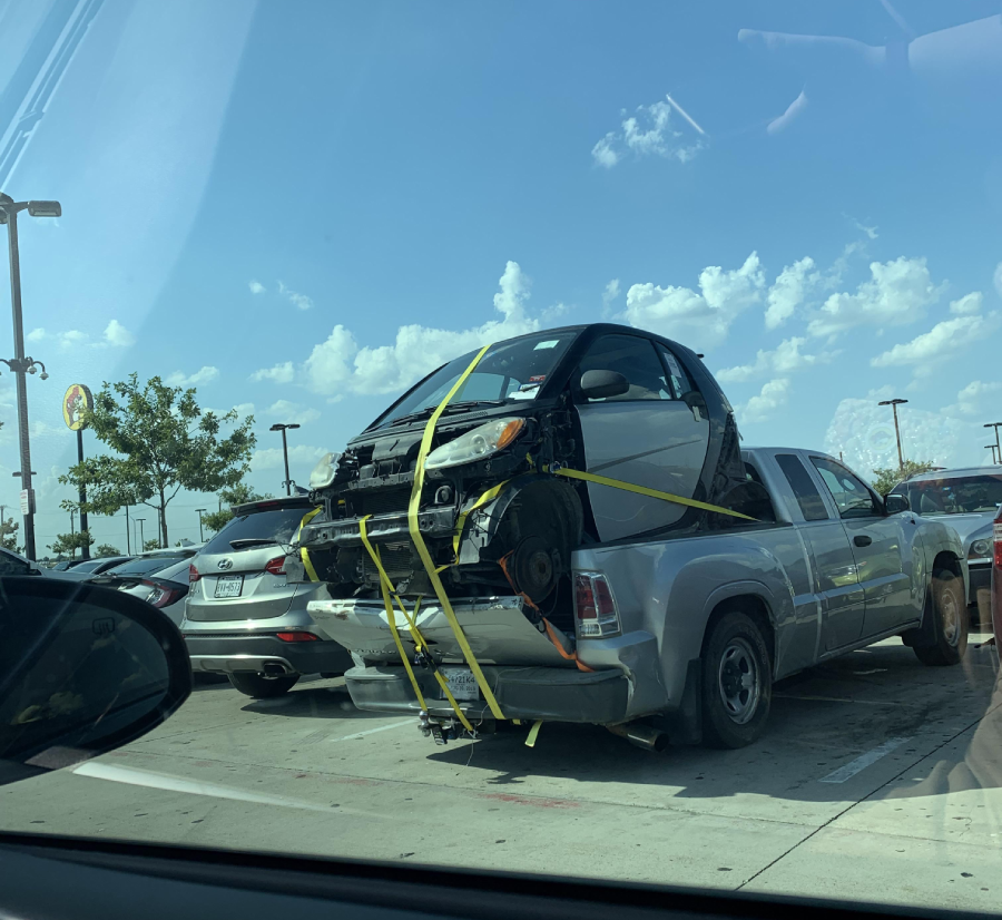 A small car missing its front and back panels is strapped onto the bed of a pickup truck in a parking lot on a sunny day, with other cars parked nearby.