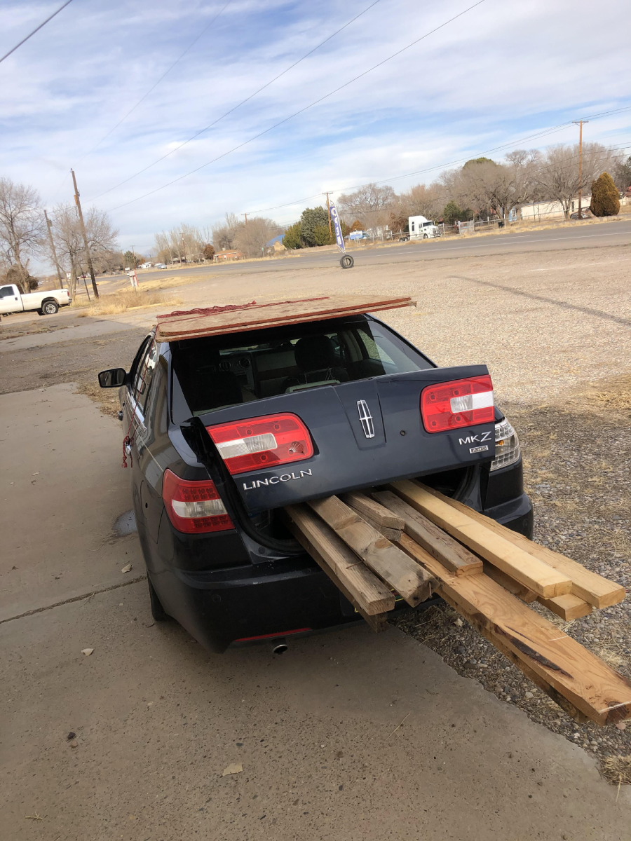 A black Lincoln sedan with its trunk open is loaded with long wooden boards sticking out from the back and some wood placed on its roof, parked on a street near a sidewalk.