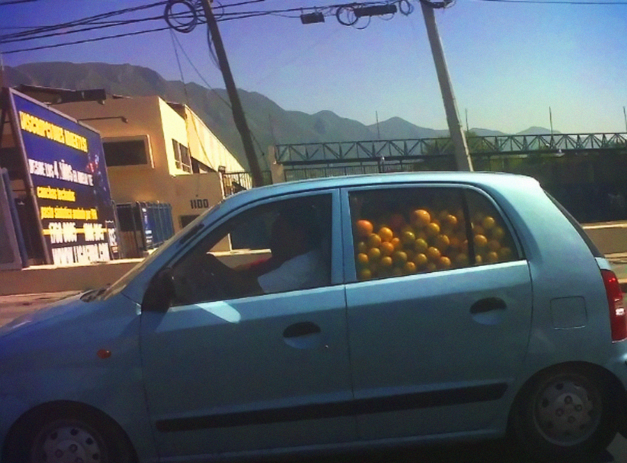 A light blue compact car with its back seat filled with oranges is driving on a sunny day, passing buildings and mountains in the background.