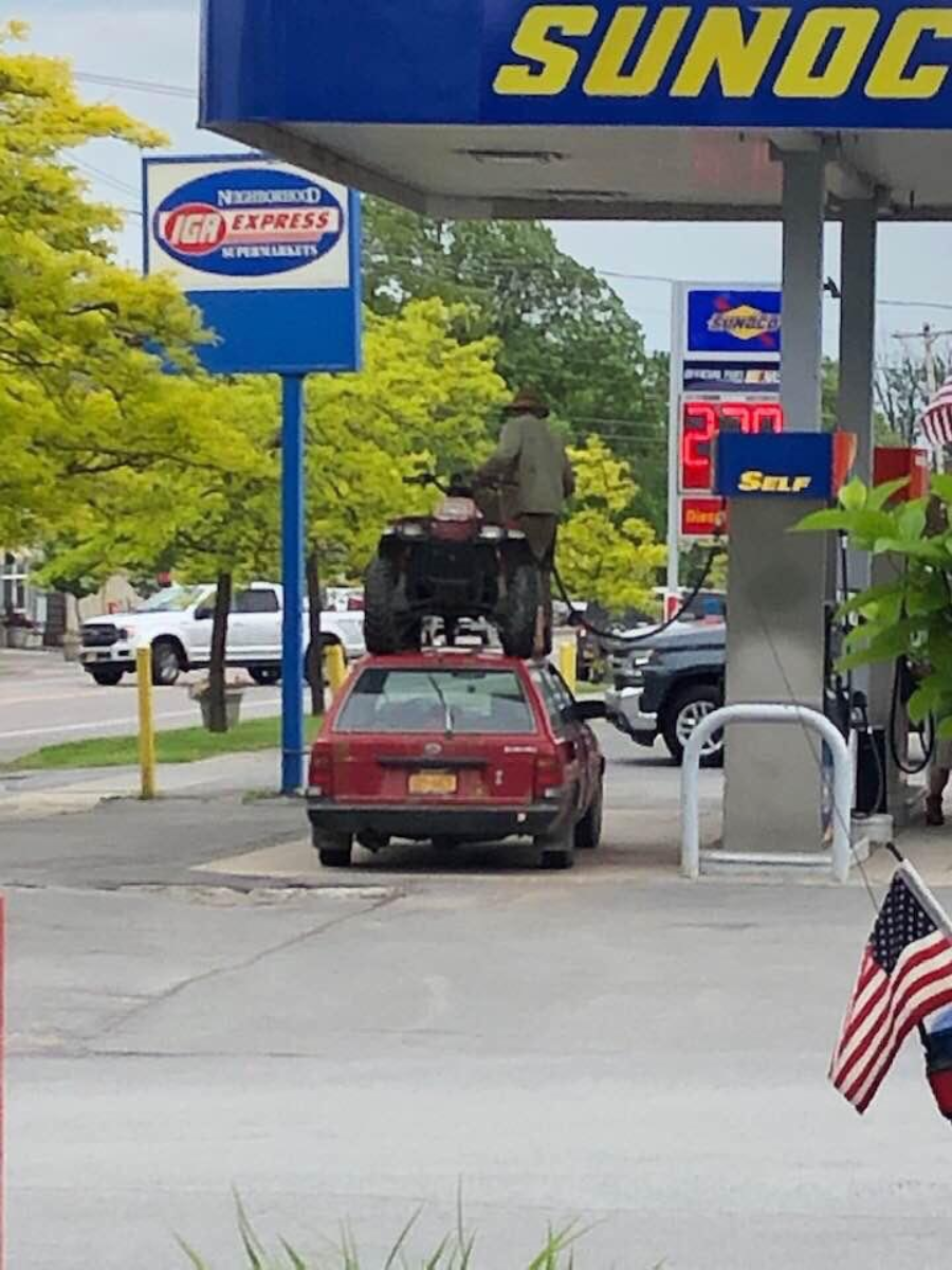 A man stands on top of an ATV that is strapped to the roof of a red car at a gas station, filling the ATV's tank at the pump. The scene is under a Sunoco canopy near an IGA Express store.