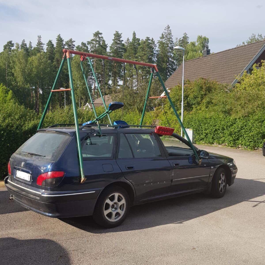 A blue station wagon is parked outdoors with a large swing set frame mounted on its roof. The frame is made of green poles with red crossbars, resembling playground equipment. Trees and houses are in the background.