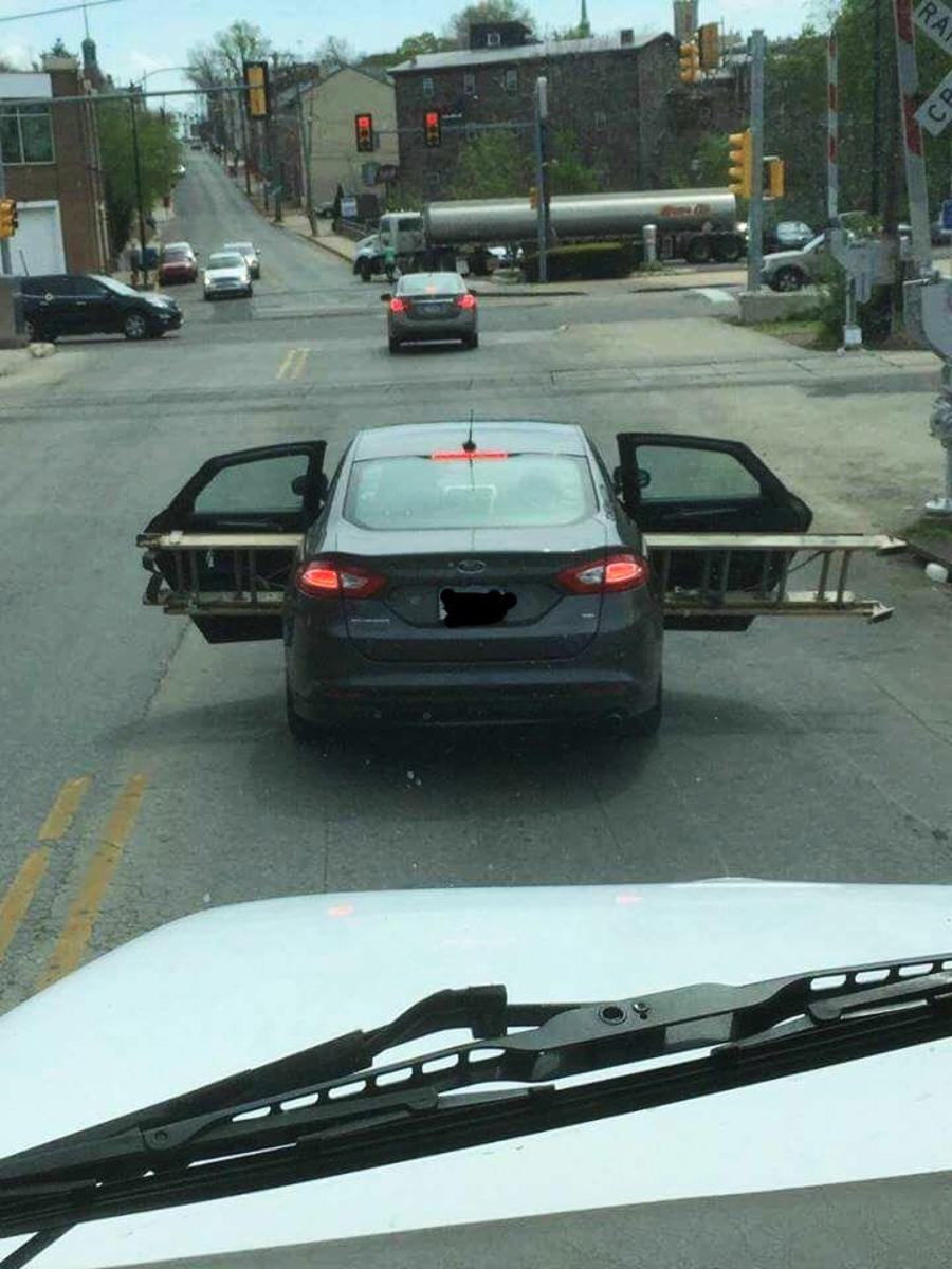 A car is driving on a city street with both rear doors wide open, each holding a long metal ladder extending out from both sides, blocking adjacent lanes.