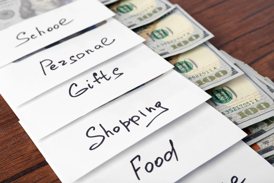 Five white envelopes labeled School, Personal, Gifts, Shopping, and Food are arranged on a wooden surface. Each envelope has cash inside, some bills partially visible, representing budgeting or saving money.