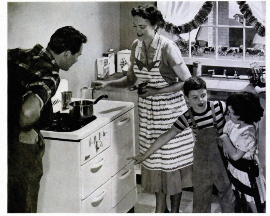 A black-and-white photo shows a family in a vintage kitchen. A man stirs a pot on the stove, while a woman and two children, all smiling, watch and point. The mood is cheerful and domestic.
