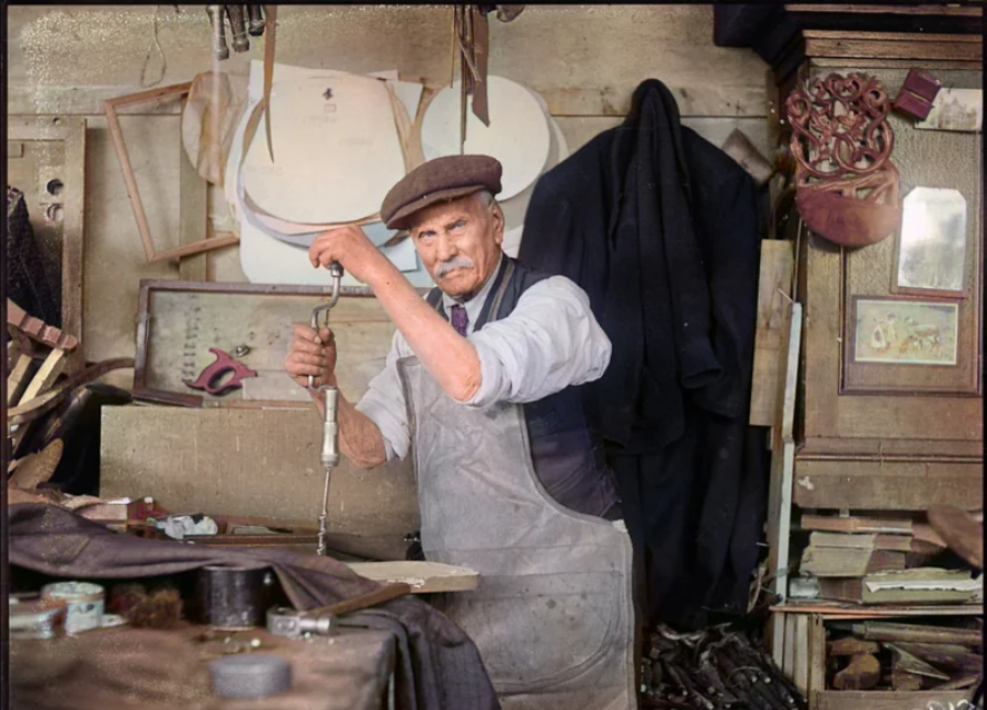 An elderly man wearing a cap and apron uses a tool at a cluttered workbench in a workshop, surrounded by various fabrics, papers, and tools hanging or stacked around him.