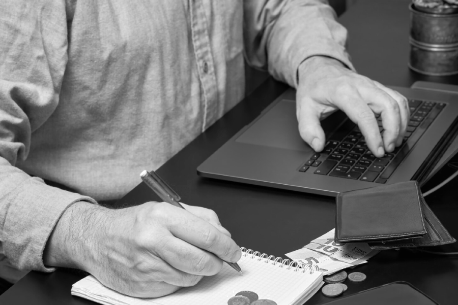 A person writes in a notebook with one hand and types on a laptop with the other; coins, a wallet, and bills are on the desk, suggesting budgeting or financial planning.