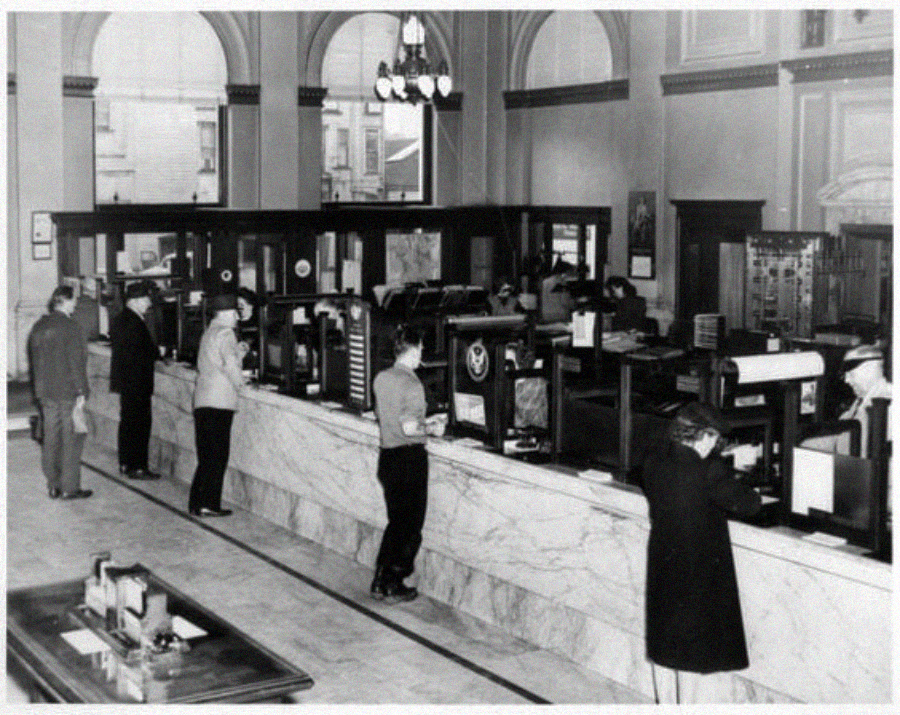 Black-and-white photo of people standing at teller windows inside a historic bank with marble counters, tall arched windows, and ornate interior details. Bank employees assist customers behind the counters.