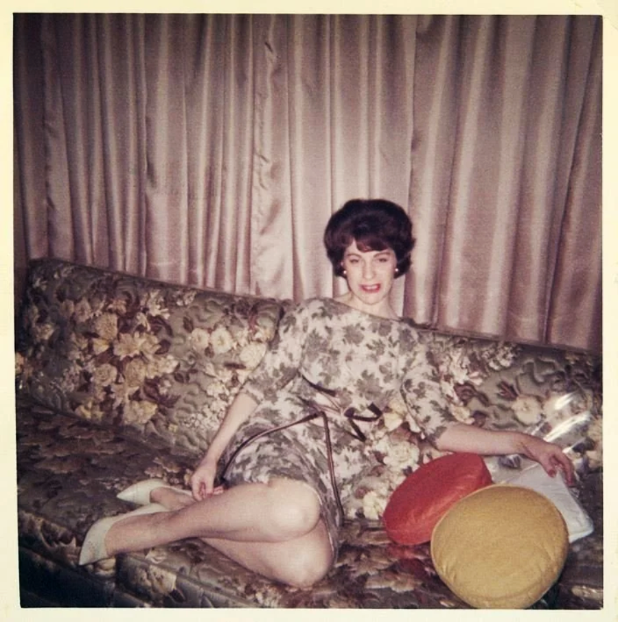 A woman with dark hair in a floral dress reclines on a floral-patterned couch. She smiles at the camera, with her legs stretched out and colorful round pillows beside her. Beige curtains hang in the background.