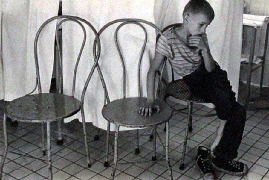 A young boy sits sideways on a metal chair, his head resting on his hand, looking thoughtful. Two empty chairs are beside him. The background features white curtains and tiled flooring.