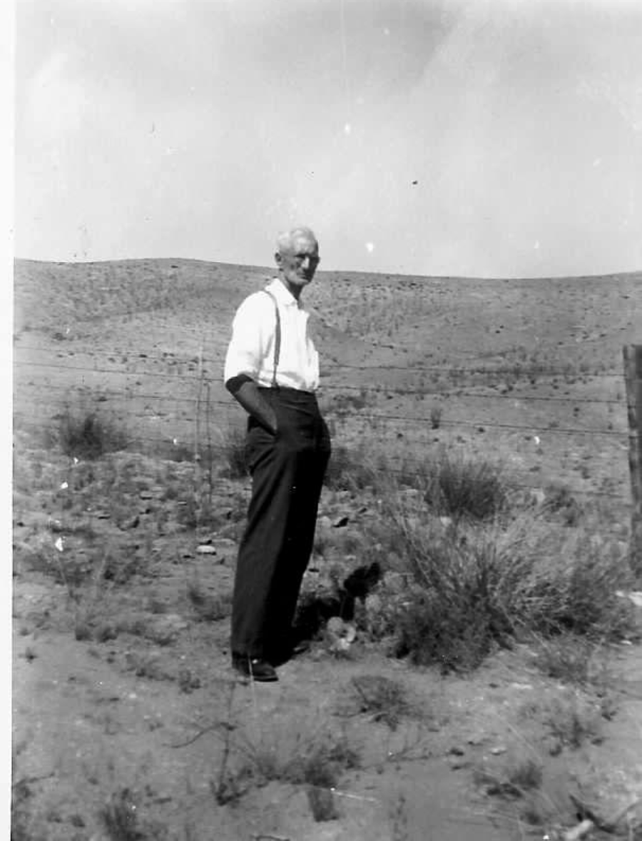 An elderly man with white hair and glasses stands outdoors in a dry, barren landscape, wearing a white shirt with rolled sleeves, suspenders, and dark trousers. There is a wire fence and hills in the background.