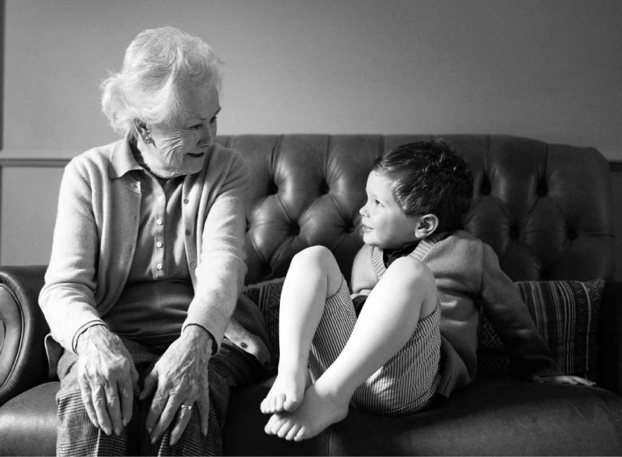 An elderly woman and a young boy sit on a couch facing each other, smiling and engaged in conversation. The boy sits barefoot with his legs up, while the woman sits with her hands in her lap. The image is in black and white.