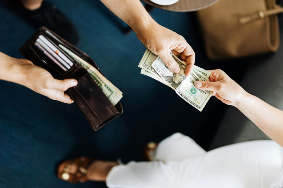 A person hands over several US dollar bills to another person holding an open wallet. The scene is viewed from above, with both individuals standing on a blue carpet.