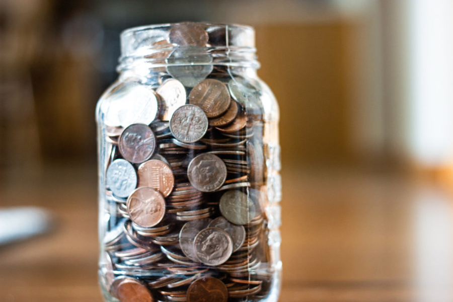 A clear glass jar filled with an assortment of coins, mostly pennies, nickels, and dimes, sits on a wooden surface with a blurred background.