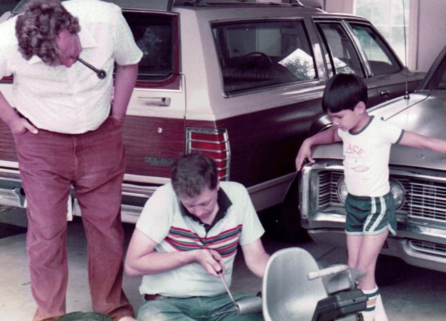 Two men and a young boy stand by a vintage station wagon in a garage. One man is working with tools, while the other man and the boy watch him closely. The boy is wearing a white t-shirt and green shorts.