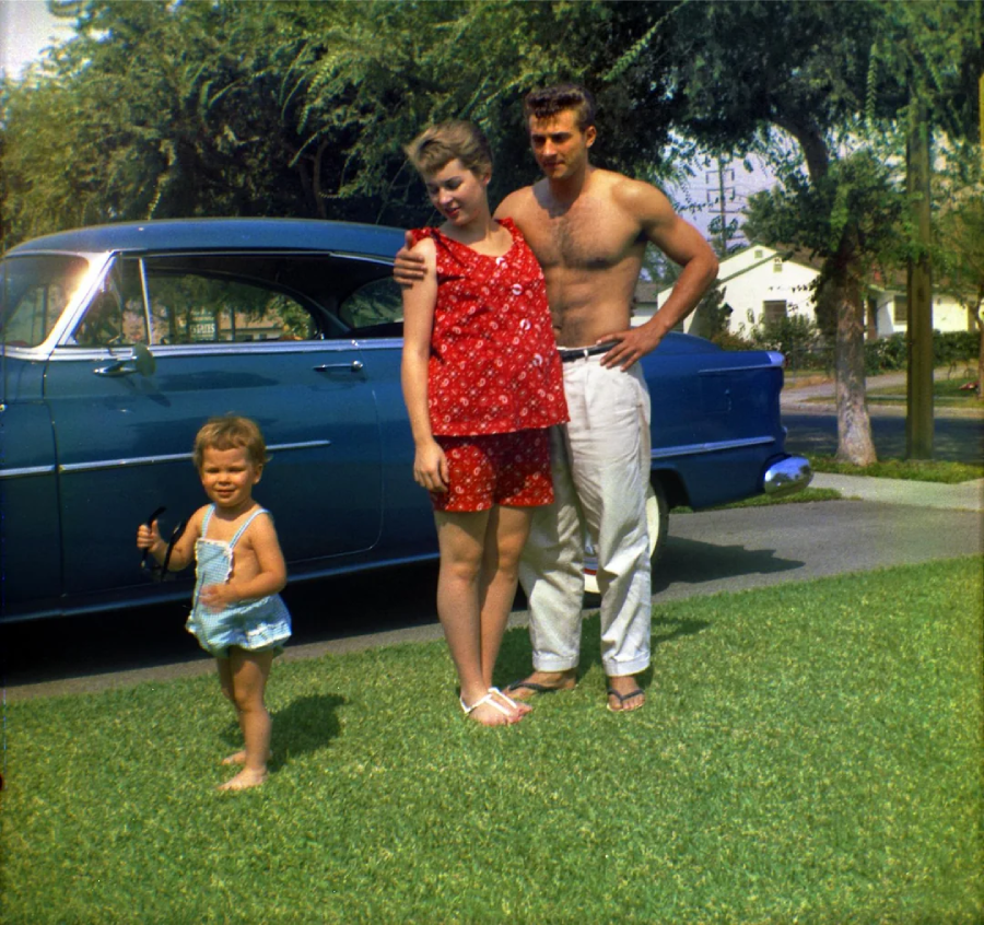 A man, woman, and young child stand on grass in front of a vintage blue car. The man is shirtless, the woman wears a red romper, and the child wears a blue outfit. Trees and houses are visible in the background.