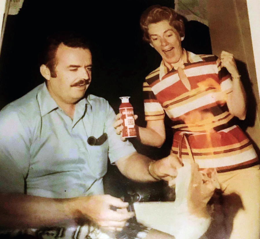 A man sits holding a pan while a woman in a striped shirt excitedly sprays something from a can, causing a burst of flames. Both appear surprised and amused.
