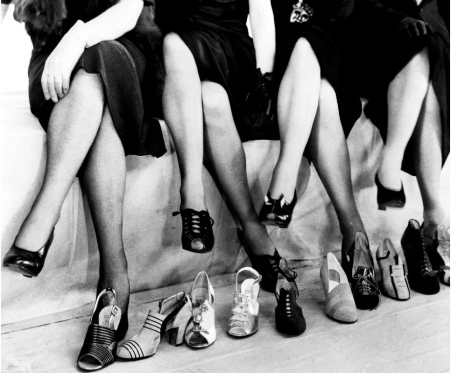 Four women in dark dresses sit with legs crossed, displaying various high-heeled shoes and sandals in front of them, showcasing a range of 1930s footwear styles.