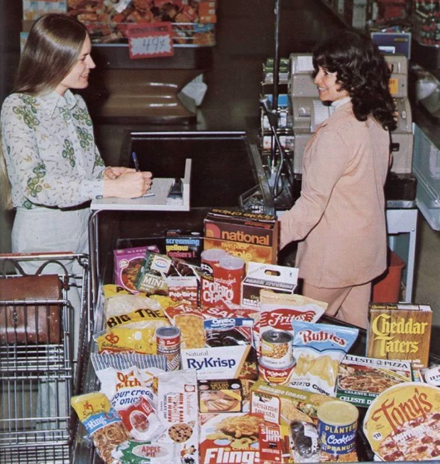 Two women are at a grocery store checkout. One woman stands behind the register, while the other stands beside a cart overflowing with colorful packaged foods and snacks, such as chips, cereal, and crackers.