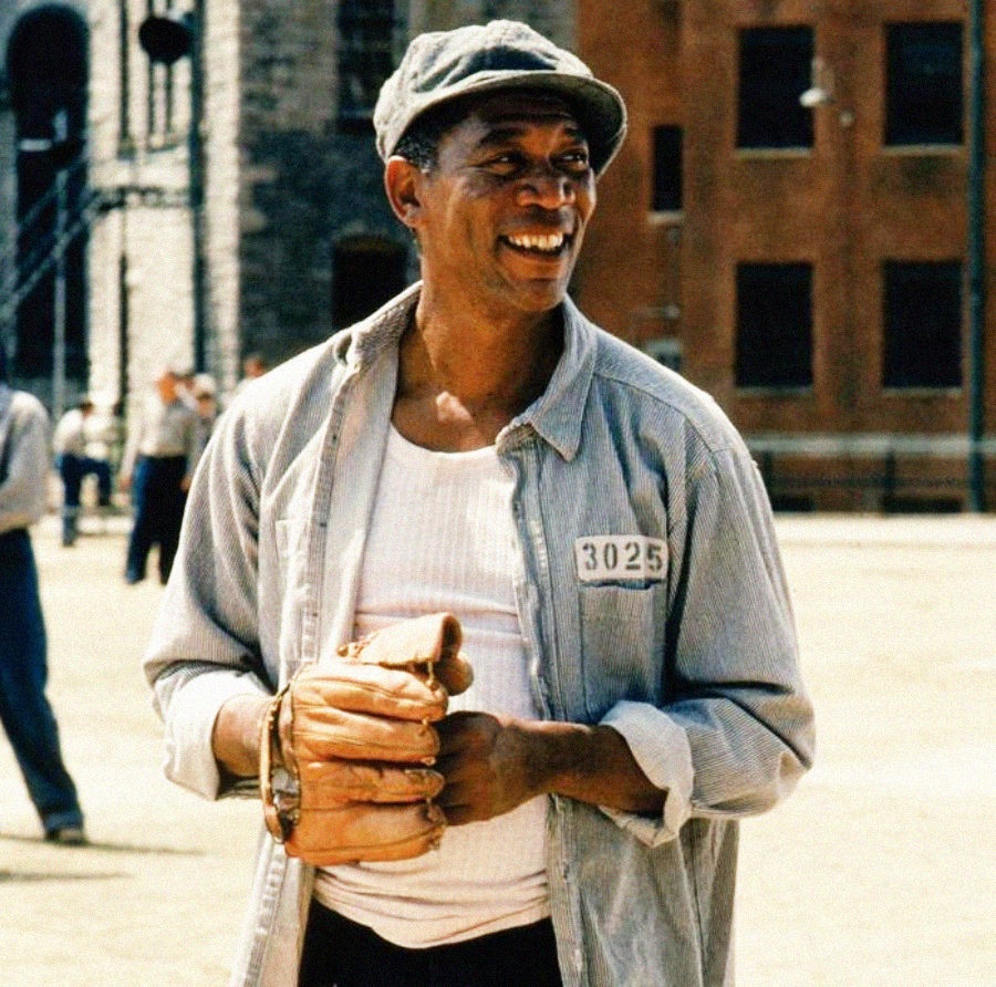 A man wearing a prison uniform, flat cap, and baseball glove stands outdoors in a prison yard, smiling. The uniform displays the number 3025, and brick buildings are visible in the background.
