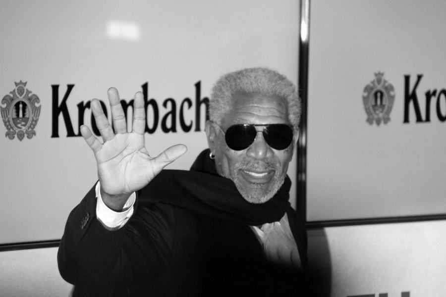 A man with gray hair and beard, wearing dark sunglasses, a scarf, and a suit, smiles and waves at the camera. The background displays a partial "Krombacher" logo. The photo is in black and white.