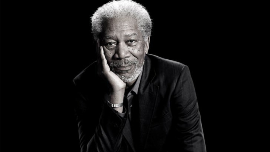 An older man with gray hair and a beard, wearing a dark suit, rests his head on his hand and looks thoughtfully at the camera against a plain black background.