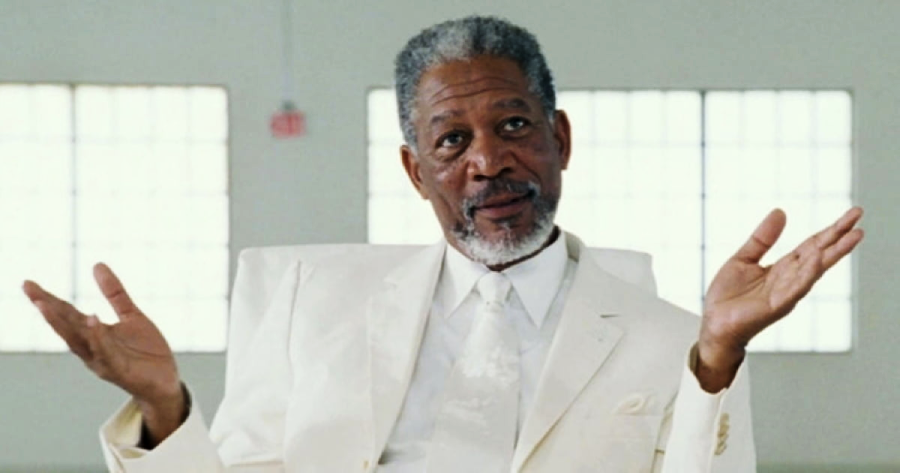 A man with gray hair and a beard, wearing a white suit, sits indoors with his hands raised and palms up, as if shrugging or explaining something, in front of large windows.
