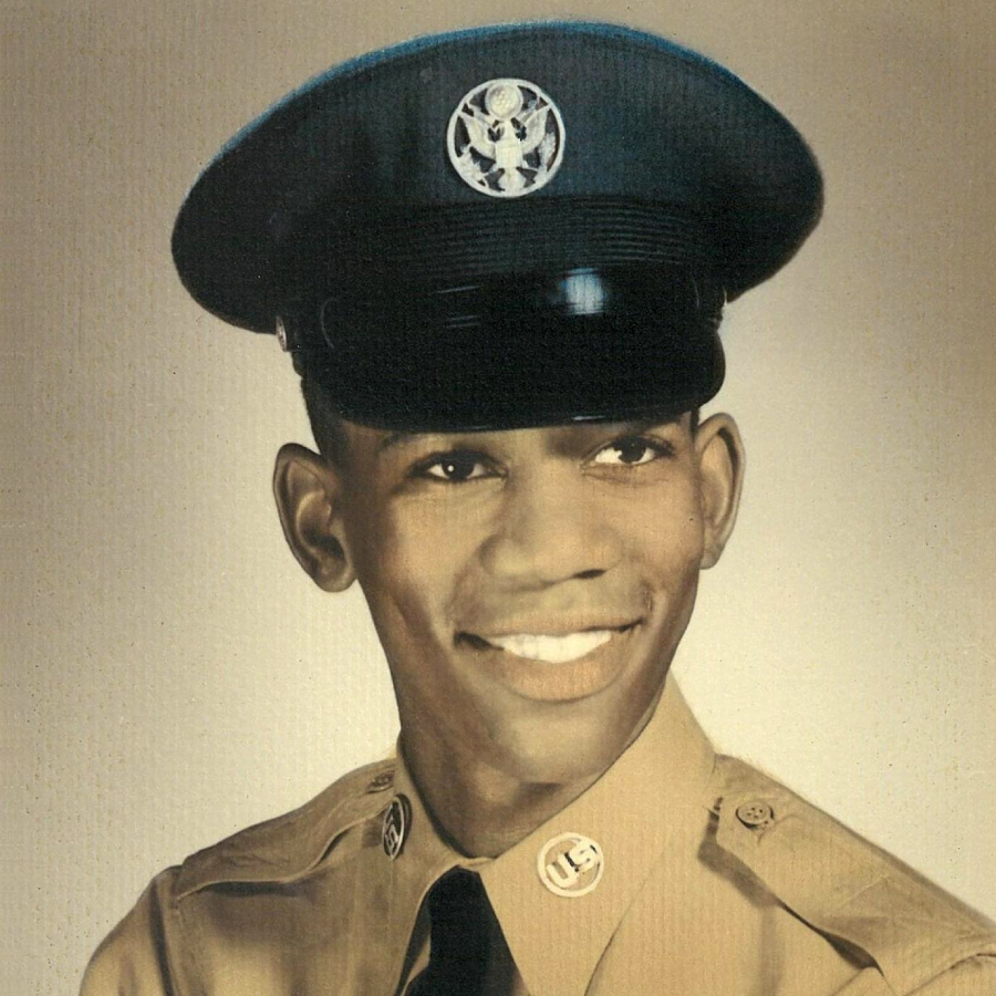 A young man in a tan military uniform and cap smiles for a studio portrait against a neutral background. He wears a black tie and has U.S. insignia on his collar and a round emblem on his hat.