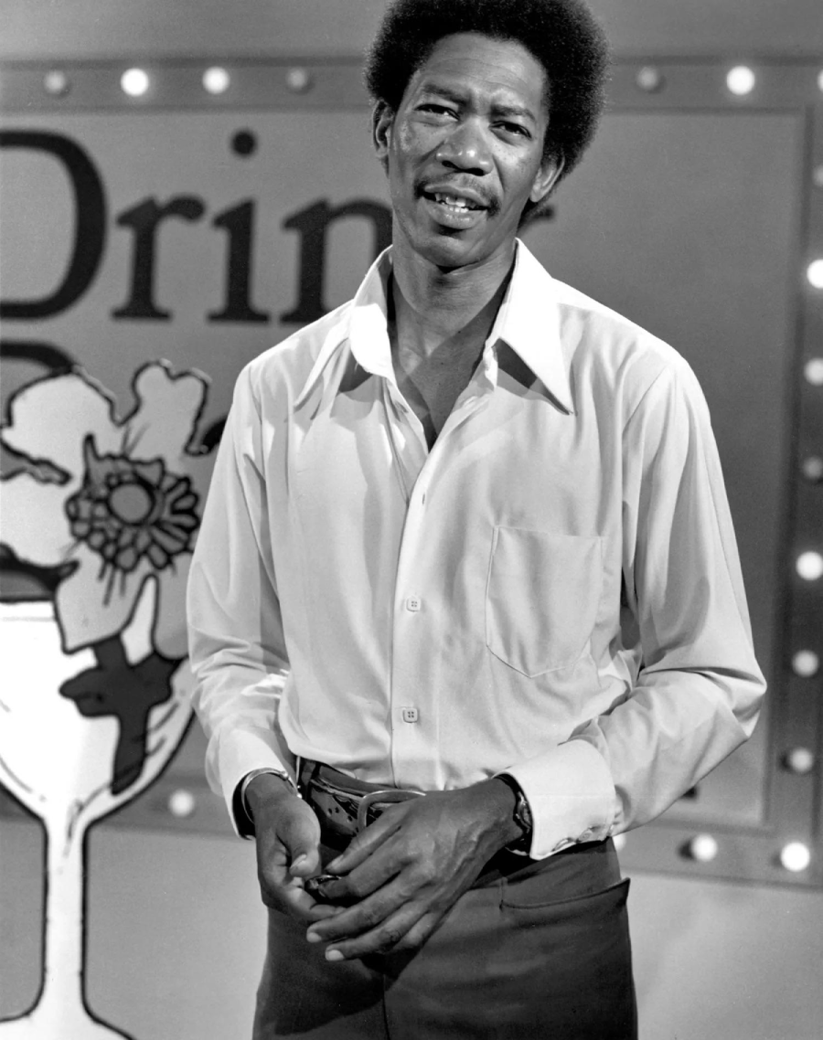 A man with an afro hairstyle wearing a light-colored button-down shirt and dark pants stands in front of a sign with a flower and a large glass, with round lights in the background.