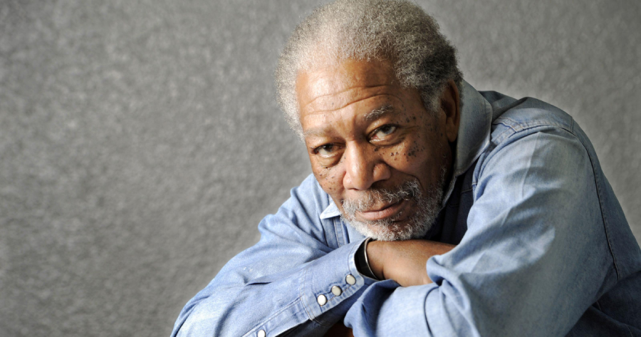 An older man with gray hair and a beard, wearing a light denim shirt, rests his arms on a surface and looks thoughtfully at the camera. The background is plain and gray.