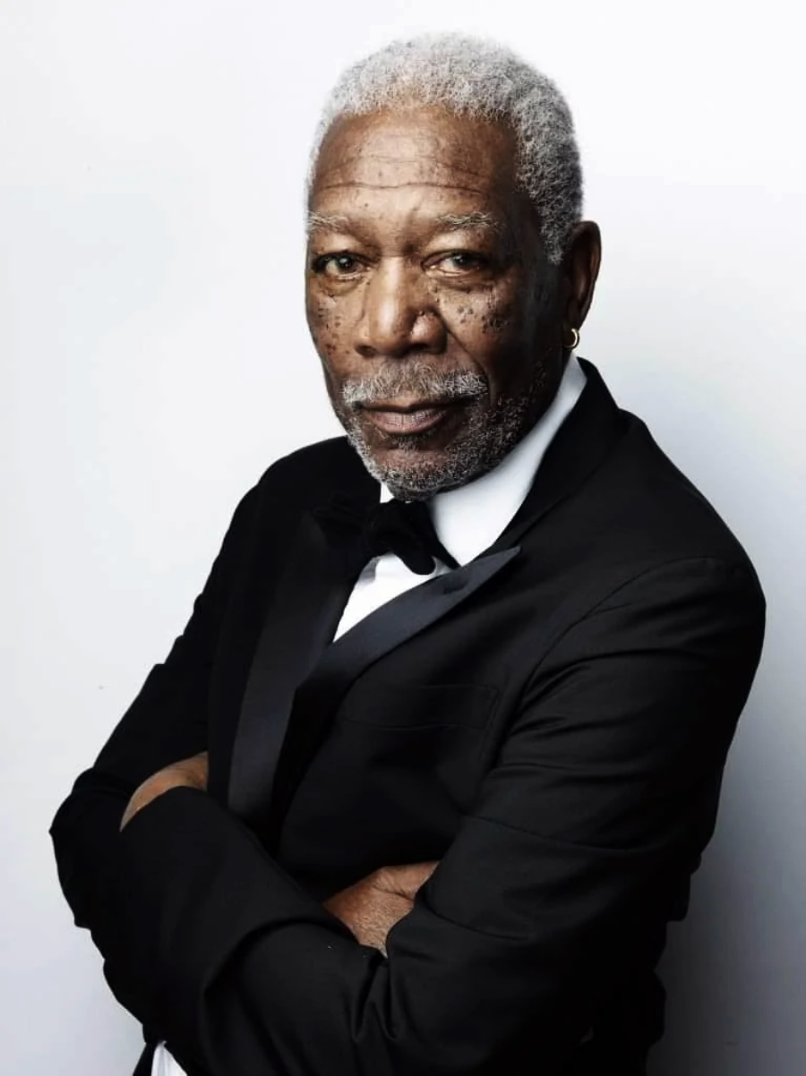 An older man with gray hair and a beard, wearing a black tuxedo and bow tie, stands against a plain white background with his arms crossed, looking confidently at the camera.