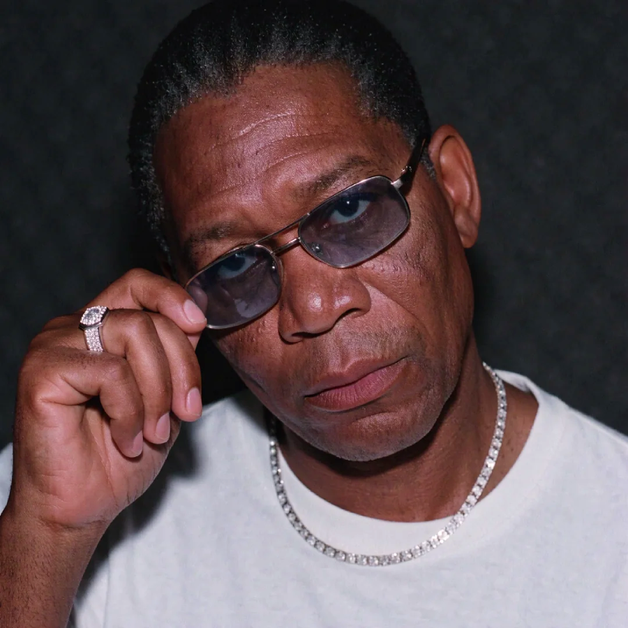 A man wearing a white shirt and silver chain looks at the camera over the top of his dark sunglasses, holding the glasses with one hand. He has short hair and wears a ring on his finger. The background is dark and textured.