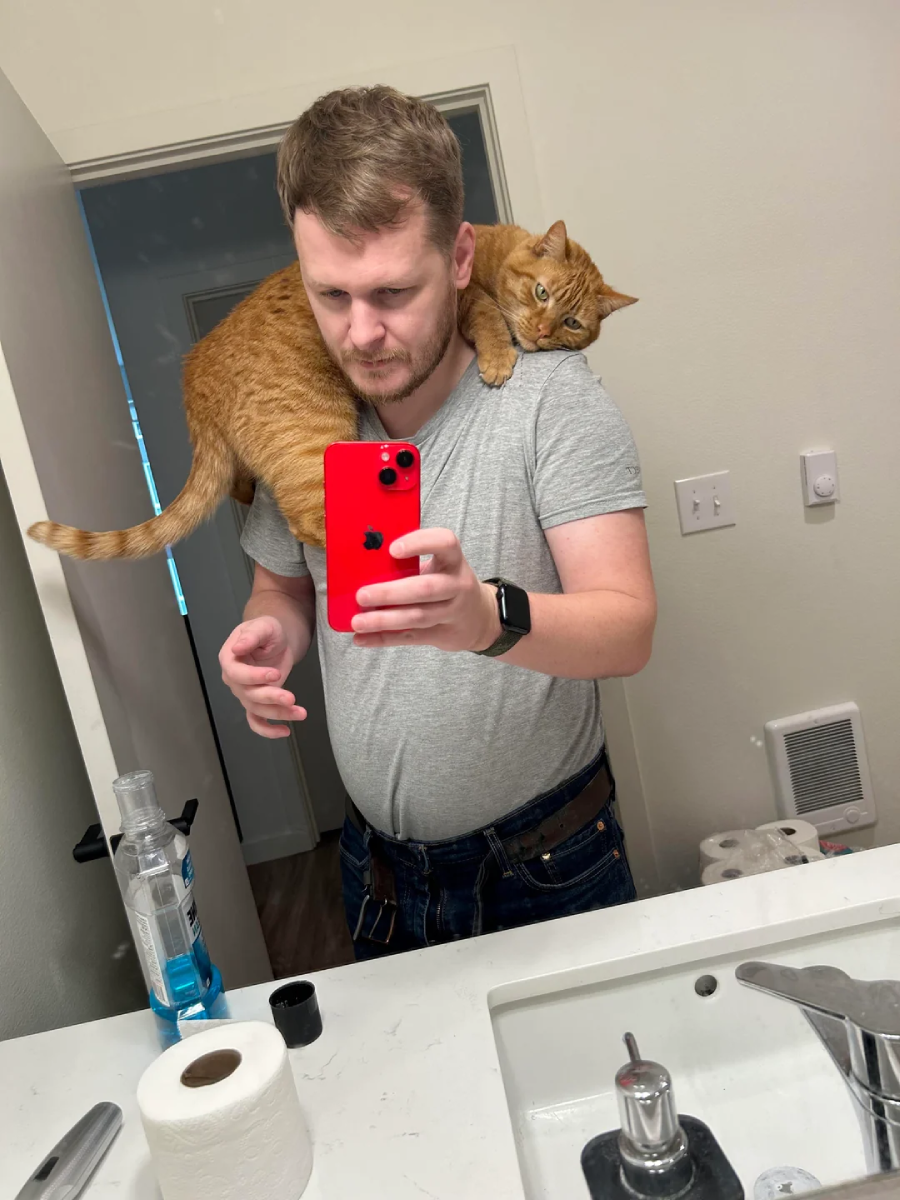 A man takes a mirror selfie in a bathroom while a ginger cat sits on his shoulders. He holds a red phone, and various items like soap and toilet paper are visible on the sink.